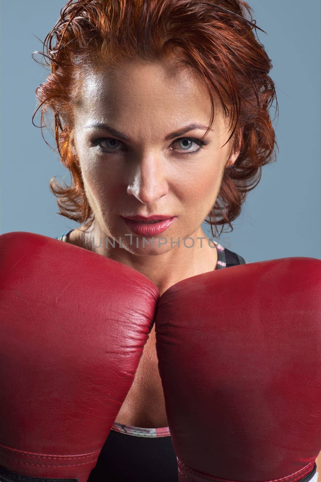Studio portrait of sporty mature woman in boxing gloves on gray background, athletic red-haired serious female looking at camera