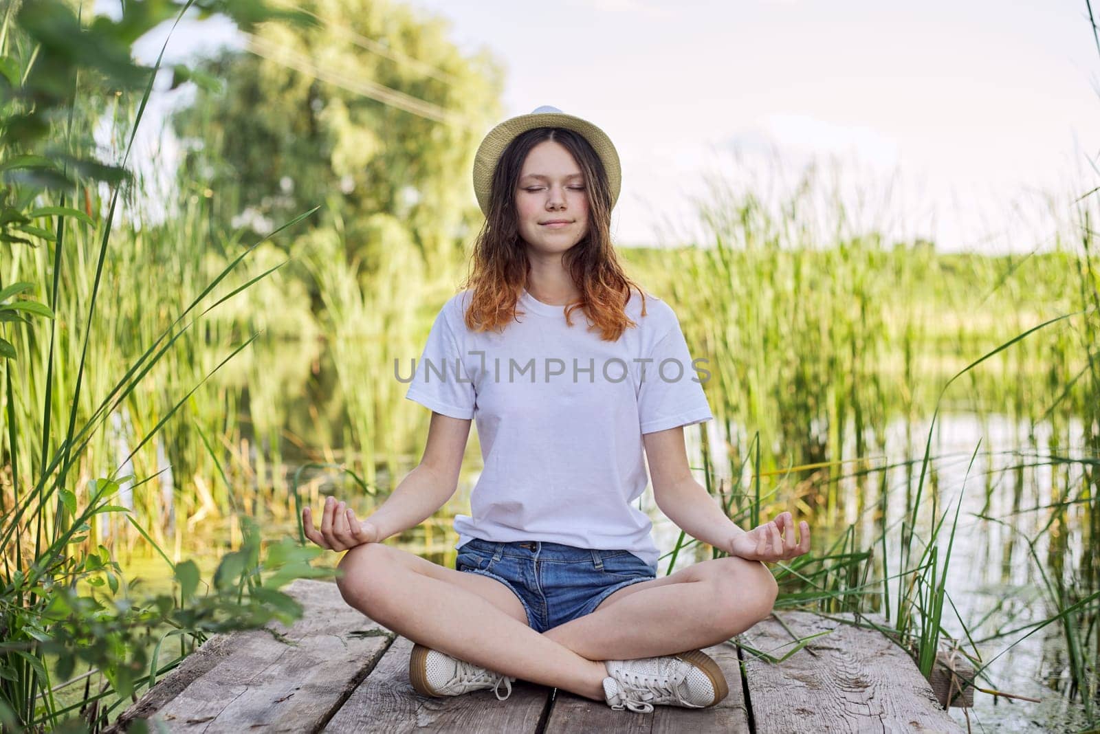 Teen girl sitting in lotus position on bridge near lake, meditating on nature