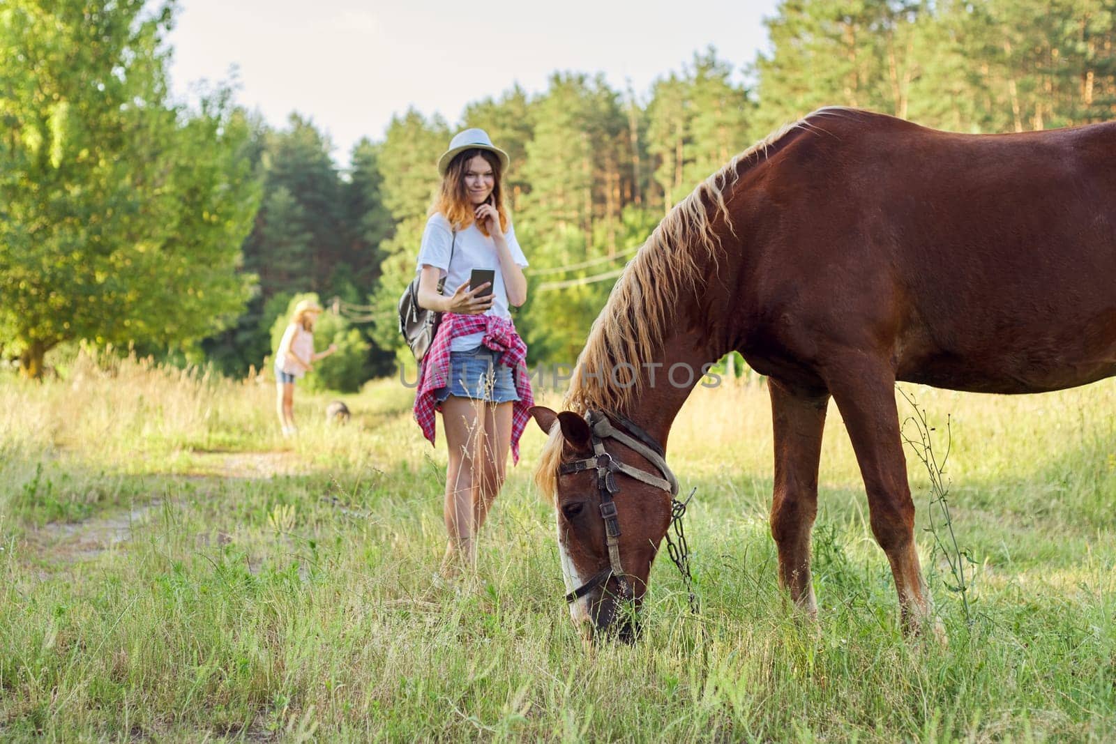 Teen girl photographing farm horse on smartphone. Rustic country style, summer nature background