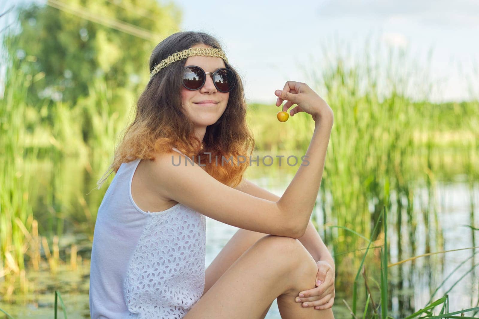 Teen girl sitting on wooden pier with yellow cherries. Beautiful young woman eating natural fruits and enjoying picturesque sunset on lake. Beauty, healthy food, nature, copy space