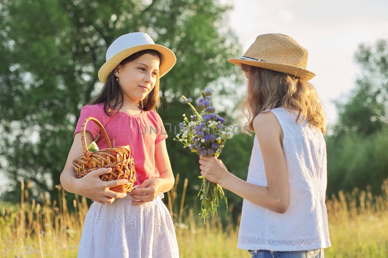 Two girls in meadow on sunny summer day, children walking, with bouquet of wildflowers basket of yellow cherries. Healthy lifestyle and food, sunset landscape background