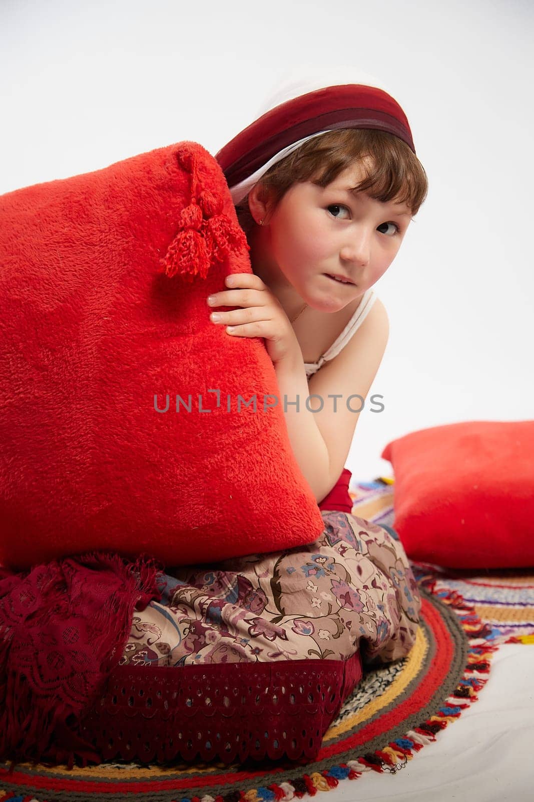 Portrait of Little girl in a stylized Tatar national costume having rest with a red pillow on a white background in the studio. Photo shoot of funny young teenager who is not professional model by keleny