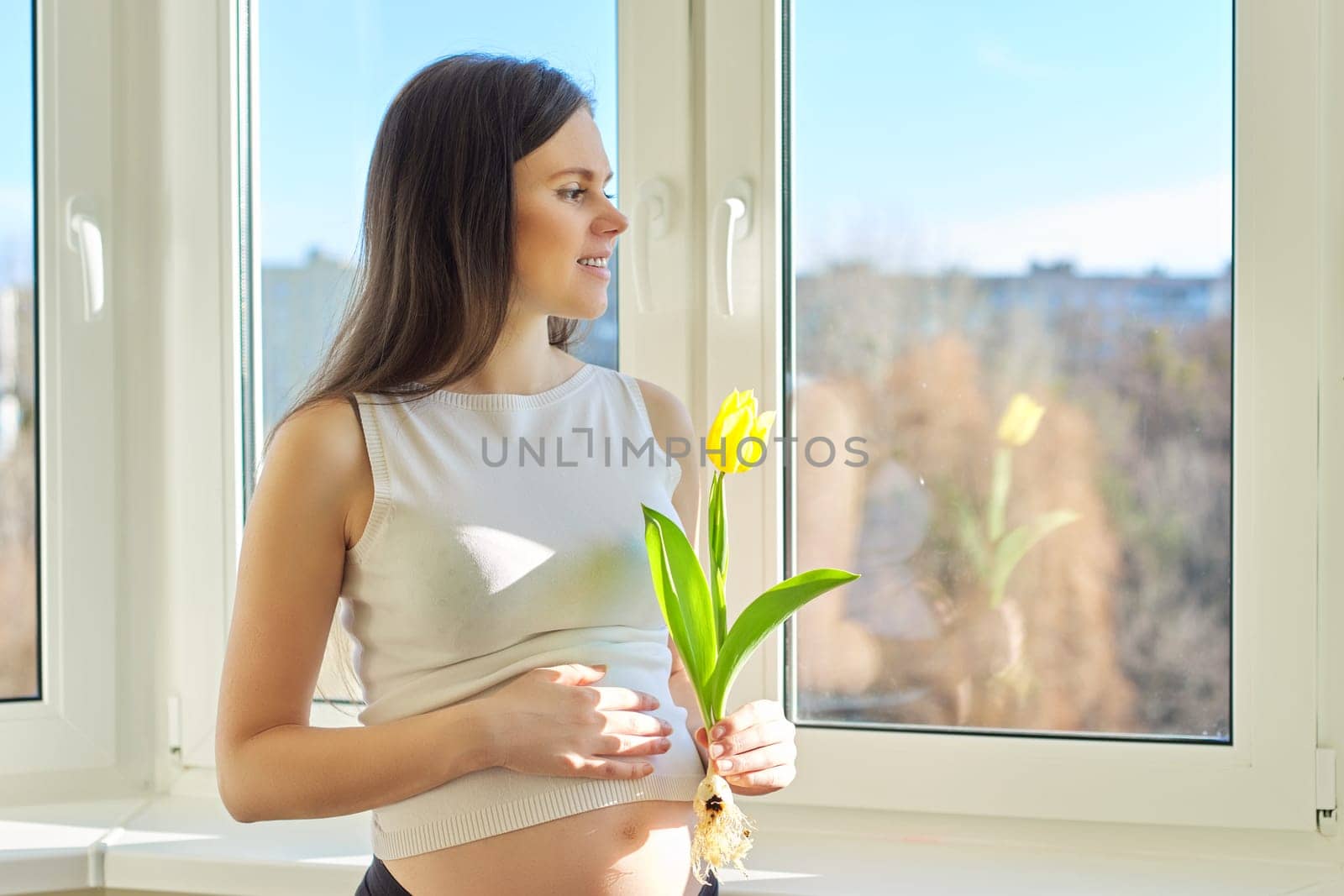 Spring, beauty, health, portrait of young pregnant woman with yellow tulip at home near window. Beautiful female with healthy skin and hair, natural makeup, copy space