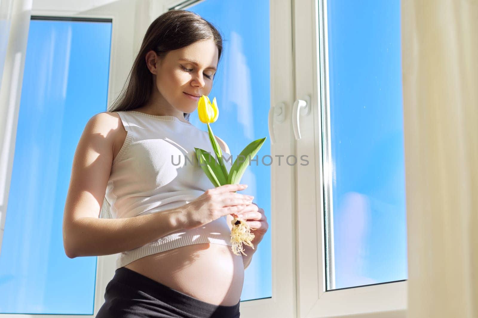 Portrait of pregnant woman with one yellow tulip flower. Spring sunny day, female in home interior near window, copy space