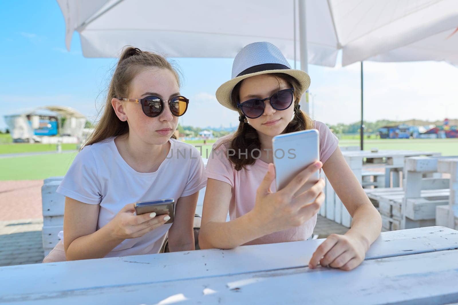 Two teenage girls with smartphones sitting and talking in summer outdoor cafe. Youth, teens, friendship, communication, people concept