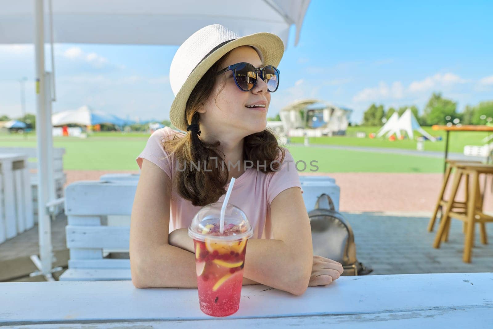 Summer fashion portrait of teenage girl in sunglasses hat sitting in outdoor cafe with refreshing berry cocktail. Vacation, rest, youth, summertime