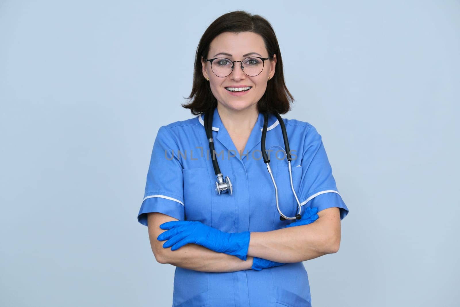 Female medical worker in blue uniform with stethoscope and gloves, confident professional woman with folded arms looking at camera, light gray background