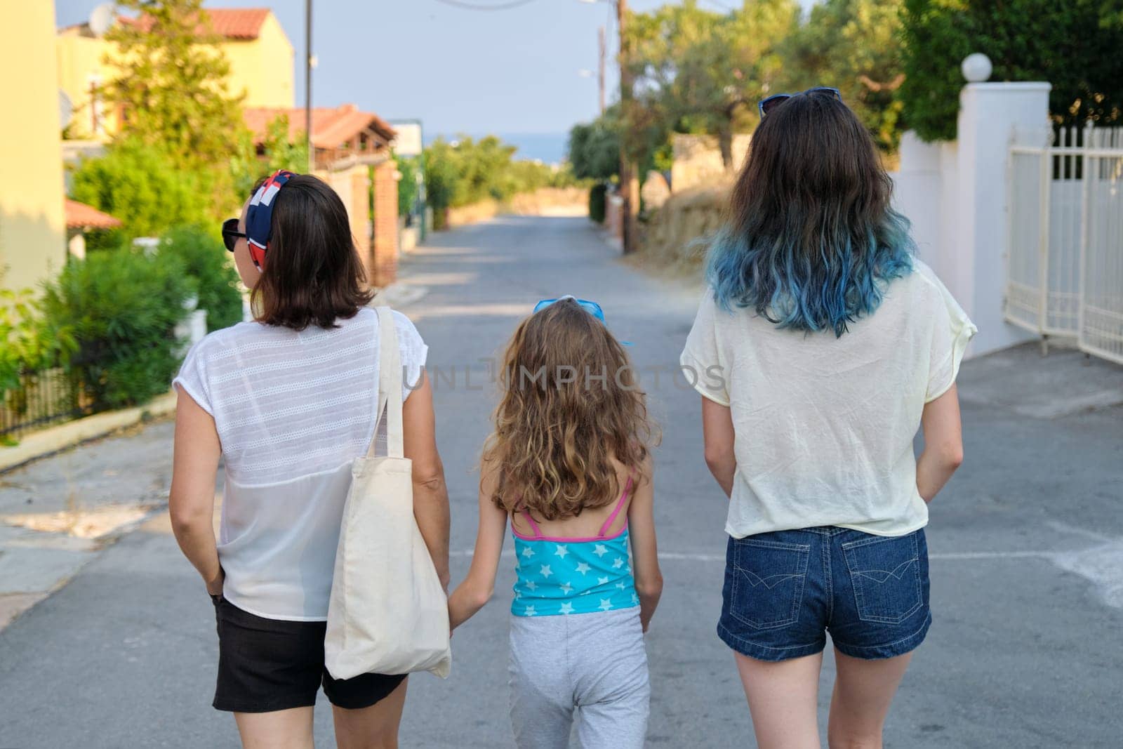 Mother and two daughters teenager and youngest walking together holding hands. Friendly family, happy parent and children, summer resort landscape background, back view