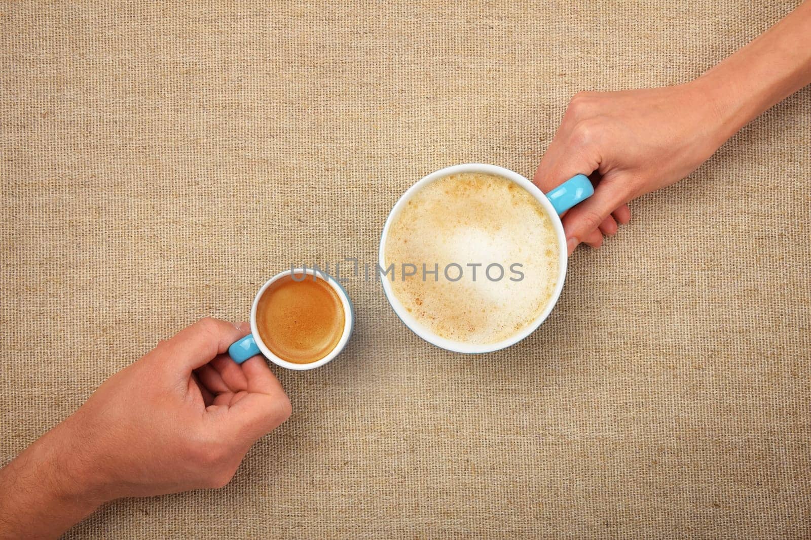 Close up man and woman hands hold two coffee cups, espresso and latte cappuccino, over burlap canvas tablecloth background, elevated, high angle view, directly above