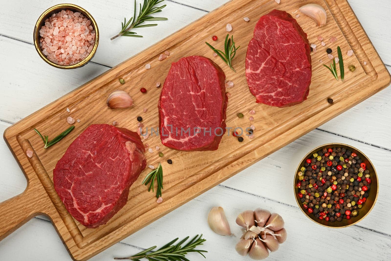 Close up three aged prime marbled raw tenderloin or fillet mignon beef steaks on brown oak wood cutting board, with spices, over white wooden table background, elevated top view, directly above