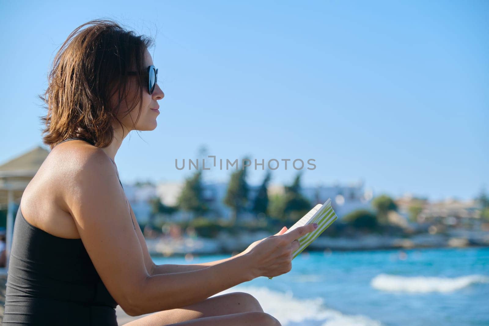 Mature woman in sunglasses swimsuit reading book sitting on sand by sea. Rest, vacation, enjoying nature, sunbathes in sunset, copy space