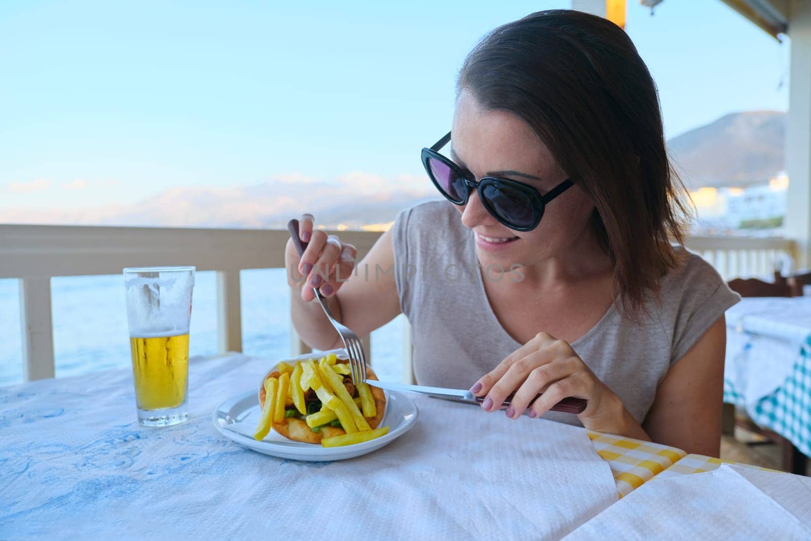 Mature beautiful woman having dinner at seaside resort cafe. Female eating and drinking light beer, enjoying meal with drink and picturesque sea sunset