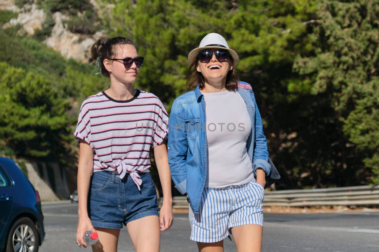 Two women mother and daughter teenager walking on sunny summer day on mountain road. Tourism, excursions, travel, family vacations