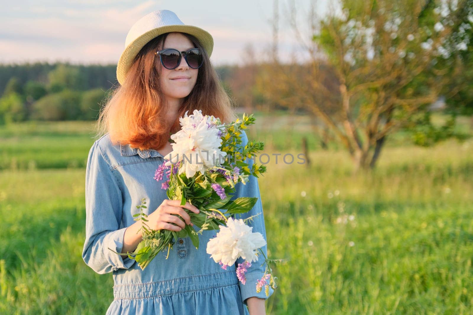 Portrait of young smiling beautiful girl in sunglasses hat with bouquet of wildflowers, sunset in meadow, copy space, rural scenery, golden hour