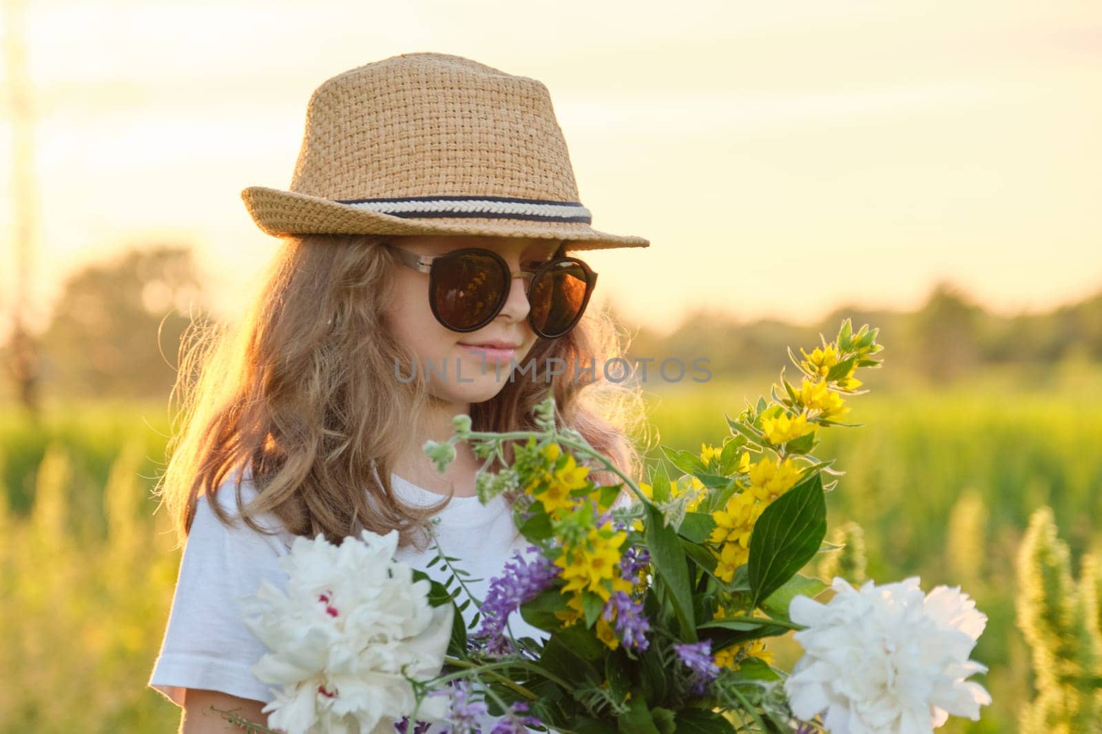 Sunny outdoor portrait of girl child 9, 10 years old in hat sunglasses with bouquet of flowers in meadow. Summer holidays, nature, childhood, beauty, golden hour