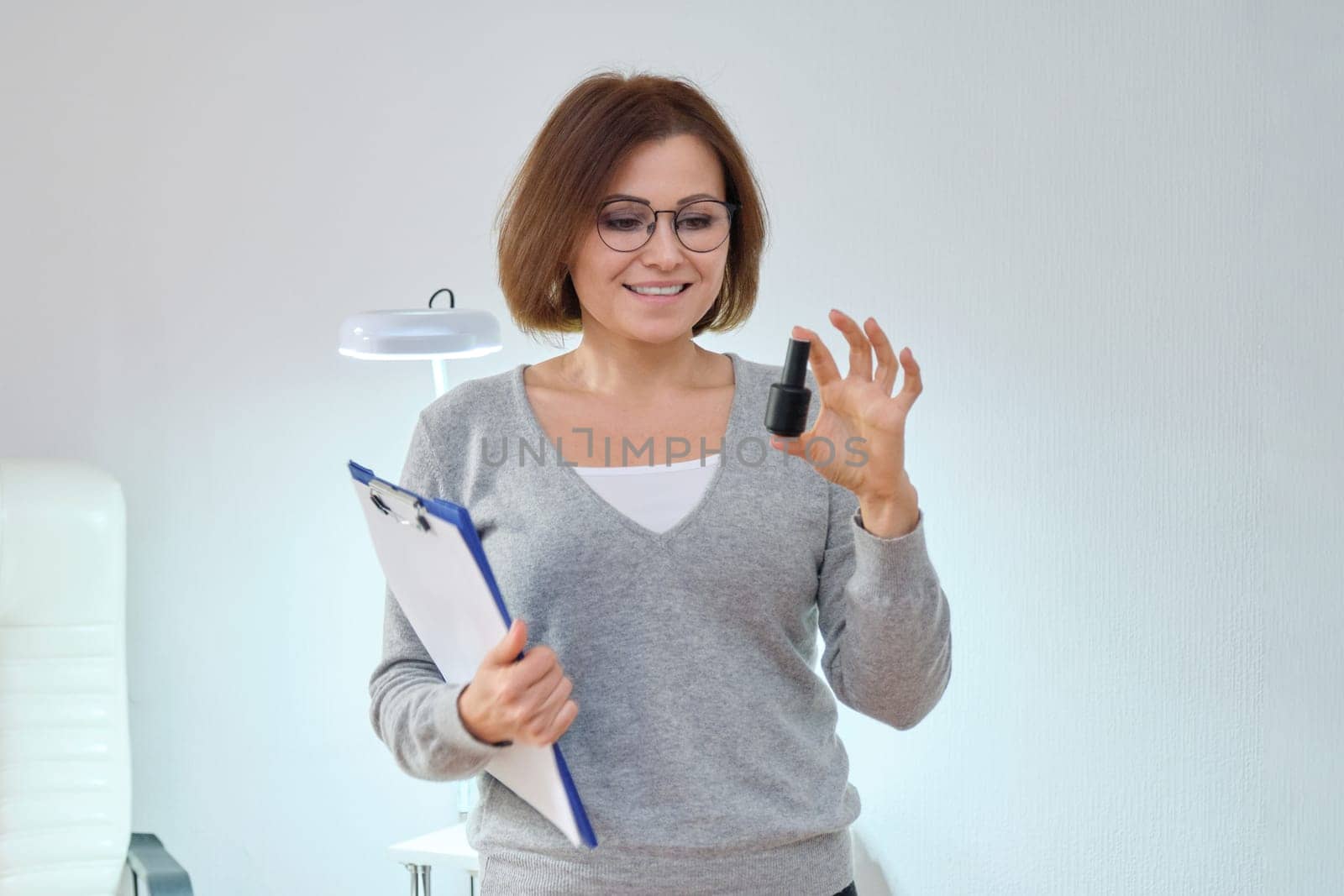 Beauty business, working mature woman owner with clipboard and nail gel polish, white interior background