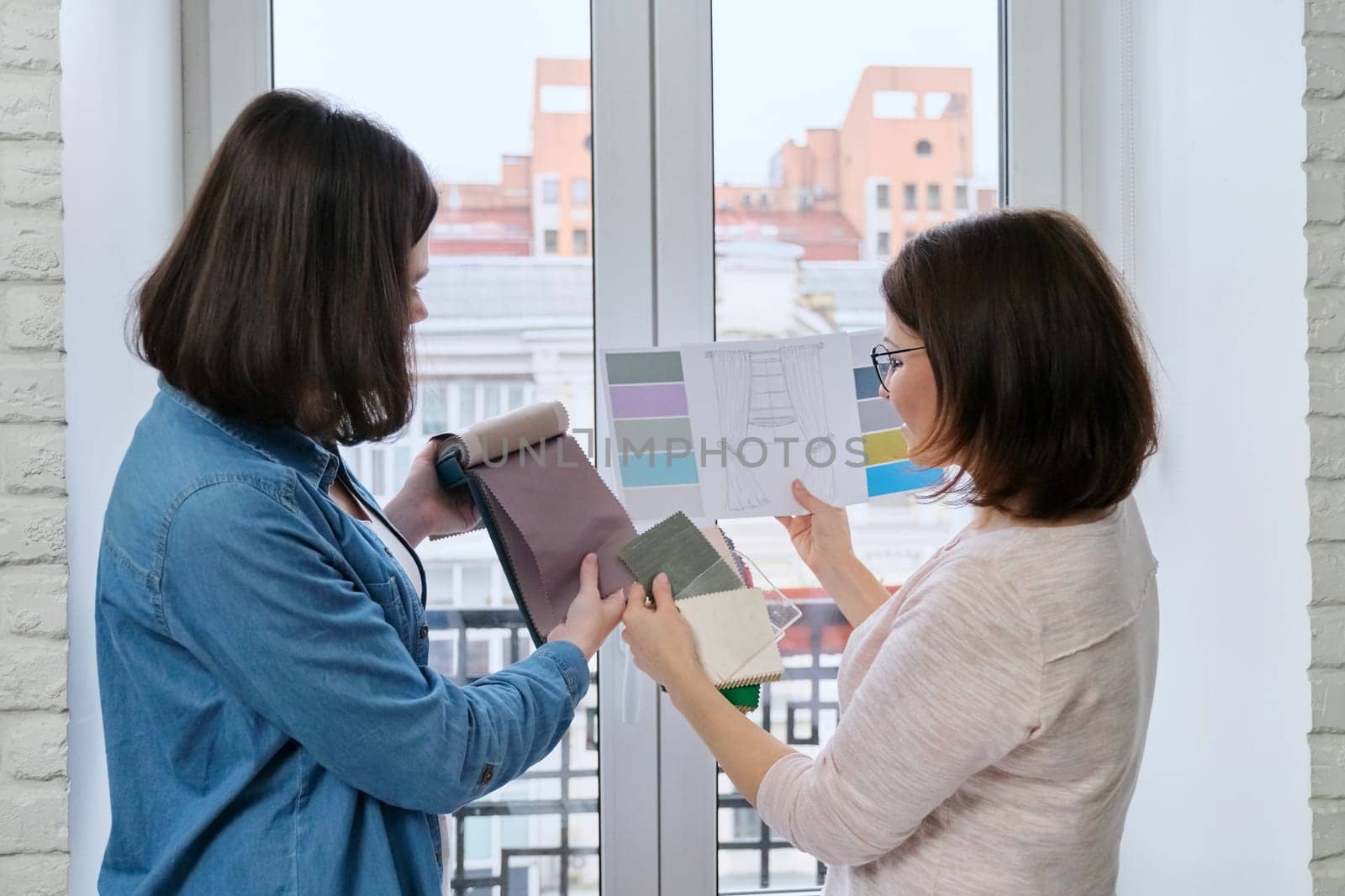 Female designer and client working with fabric samples. Selecting fabrics and design of curtains, females near window with sketch and materials