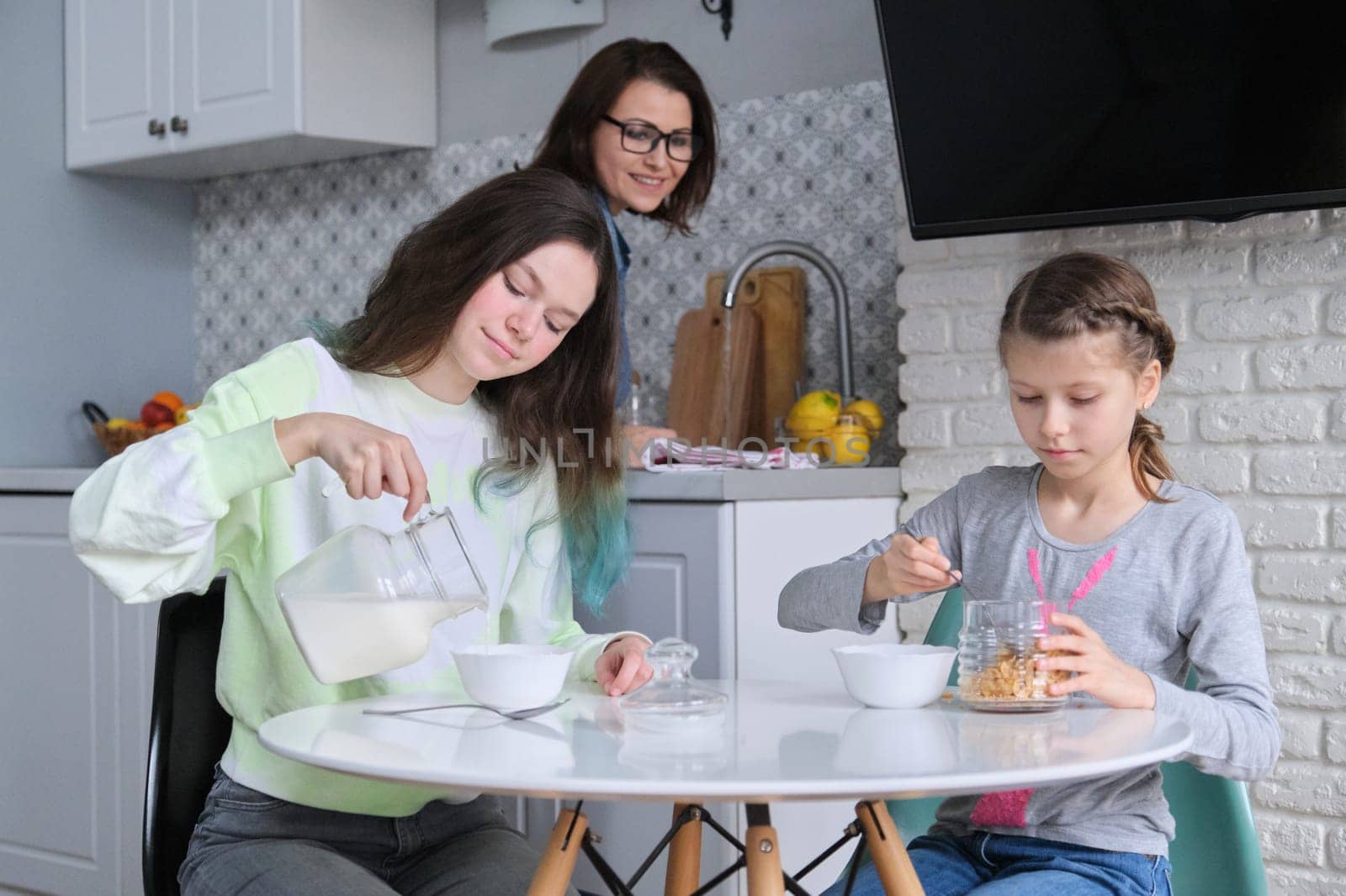 Girls having breakfast sitting at table in home kitchen, teenage sisters and 9, 10 year old child eating cornflakes with milk together. Family, food, communication, health concept.