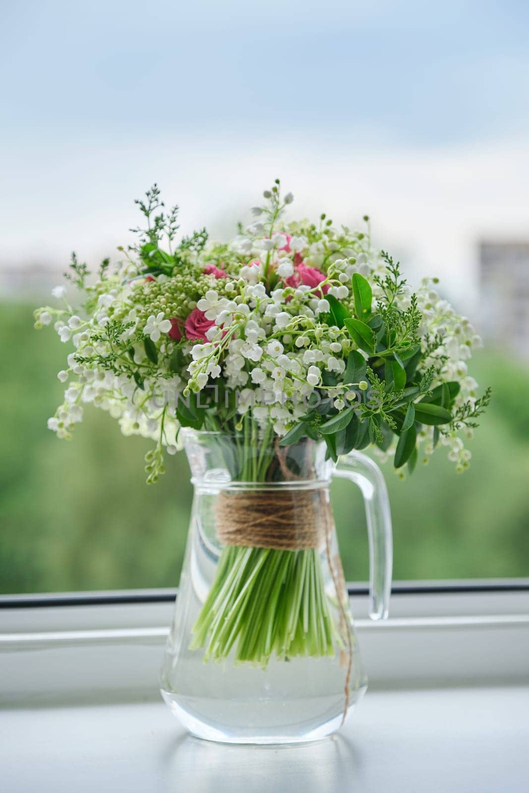 Springtime, spring fresh bouquet of lilies of the valley, pink roses, blooming viburnum. Bouquet in glass jug on windowsill, open window background