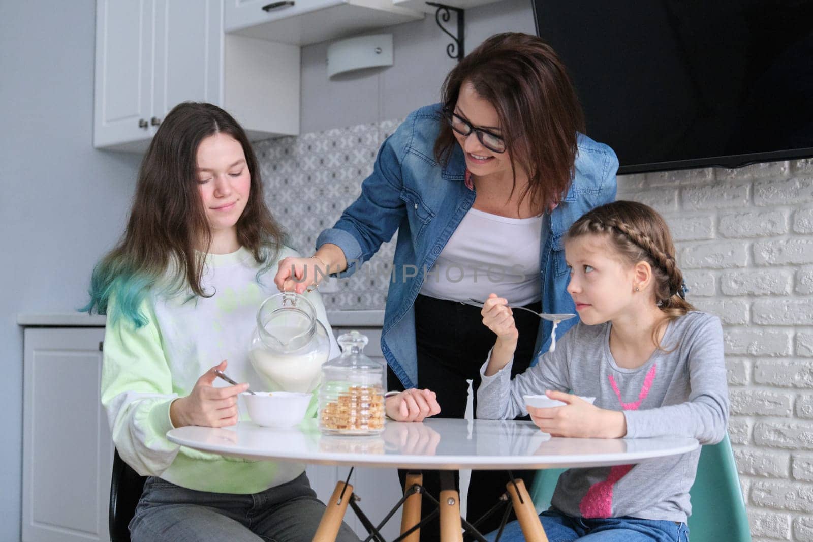 Children with mother eating at home in kitchen, two girls sitting at table with plates of corn flakes and milk. Family, eating, communication, health concept.