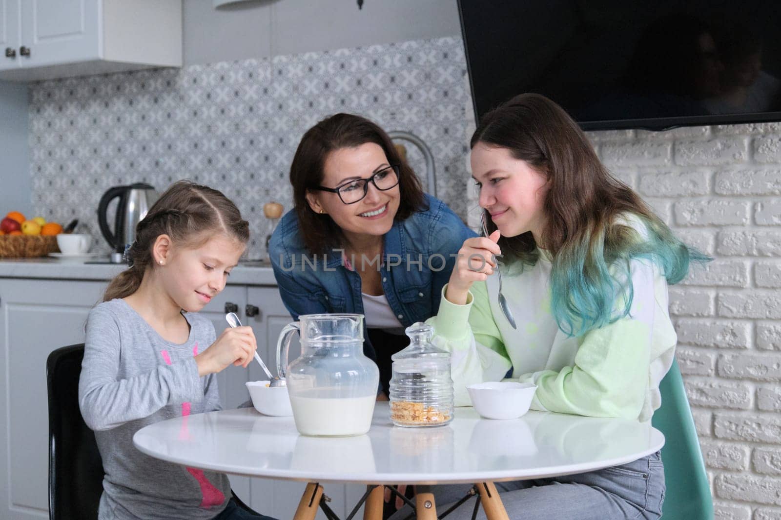 Happy mother caring for daughters in kitchen, eating girls sitting at table by VH-studio