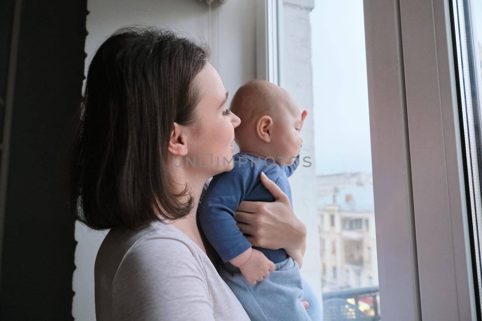 Young woman with little baby son in her arms. Mother and baby stand at home near window, look out the window