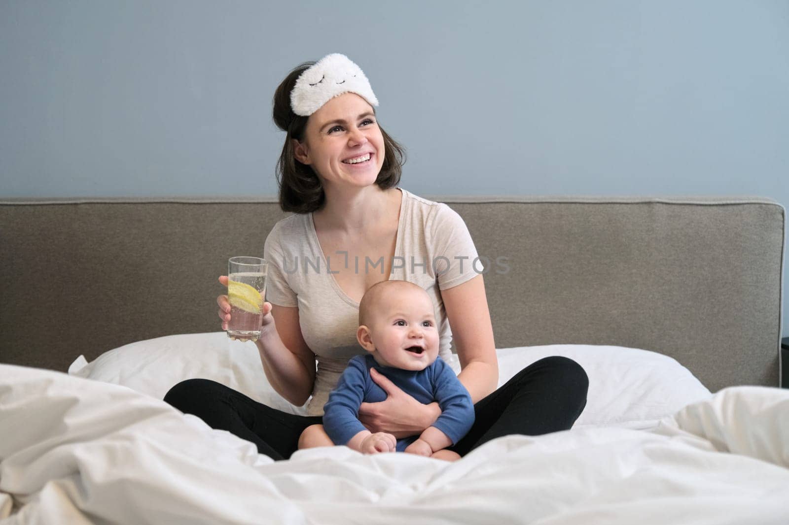 Young woman with baby toddler in arms drinking clear water with slice of lemon, sitting at home in bed. Healthy food drink for mother and baby