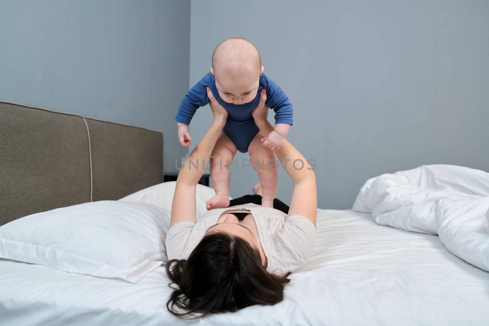 Young mother playing with toddler son, smiling woman holding baby in arms, parent and baby together at home lying in bed