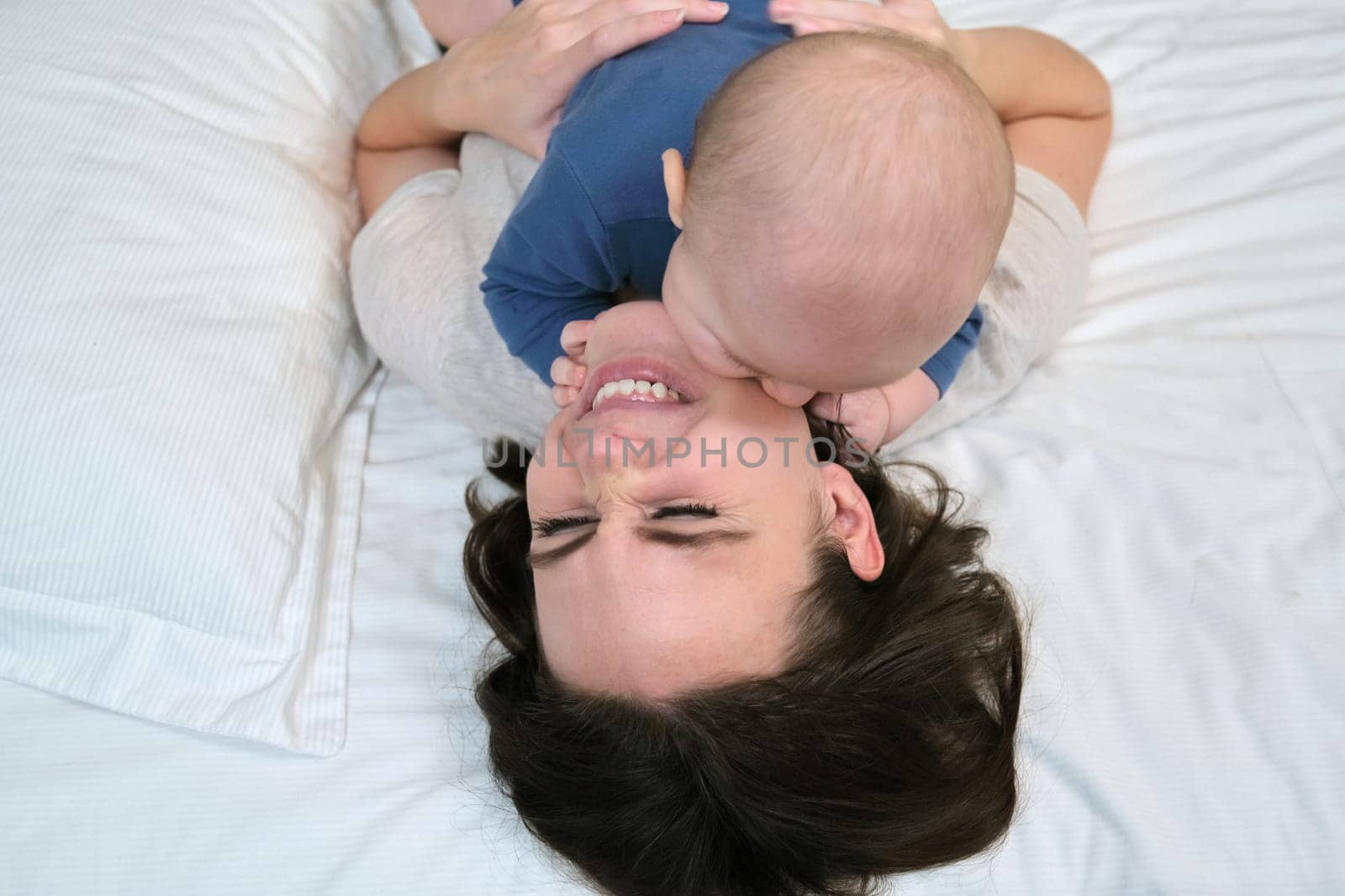 Young mother playing with toddler son, smiling woman holding baby in arms, parent and baby together at home lying in bed, view from above