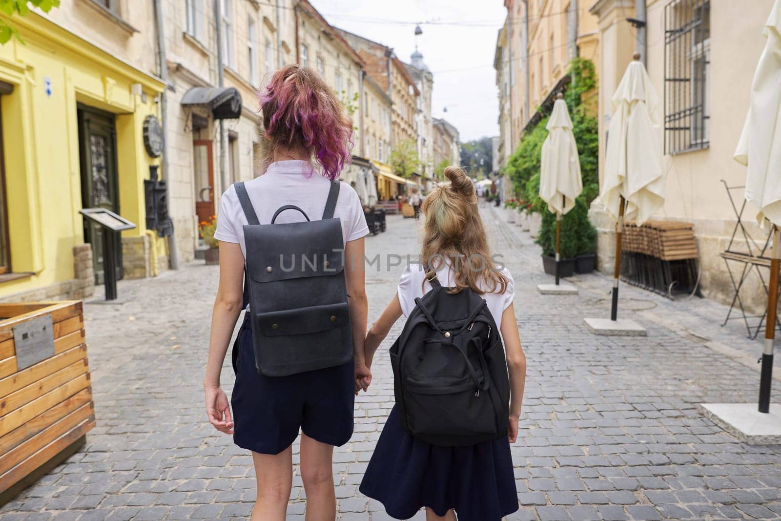 Children going to school, two girls sisters teenager and elementary school student with backpacks walking together, holding hands, back view