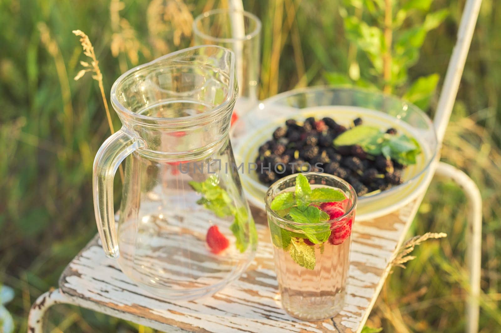 Summer natural vitamin berry drinks, jug and glasses with drink strawberry mint, plate with mulberry, background of summer herbs flowers on lawn, sunny day