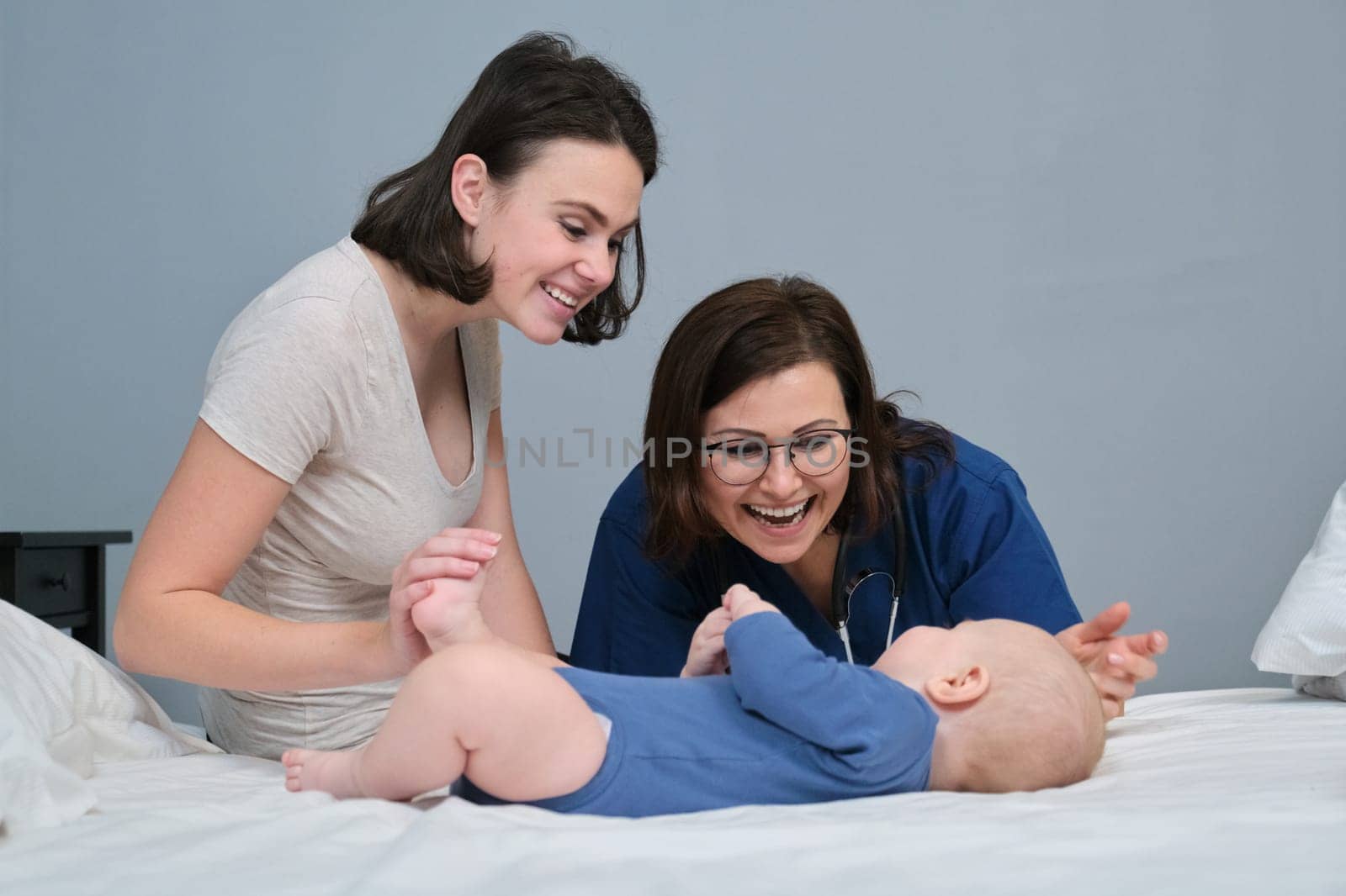 Pediatrician woman doctor in blue uniform with stethoscope talking to young mother of little boy, examining 7 month old baby at home. Pediatrics, care and health of children up to one year