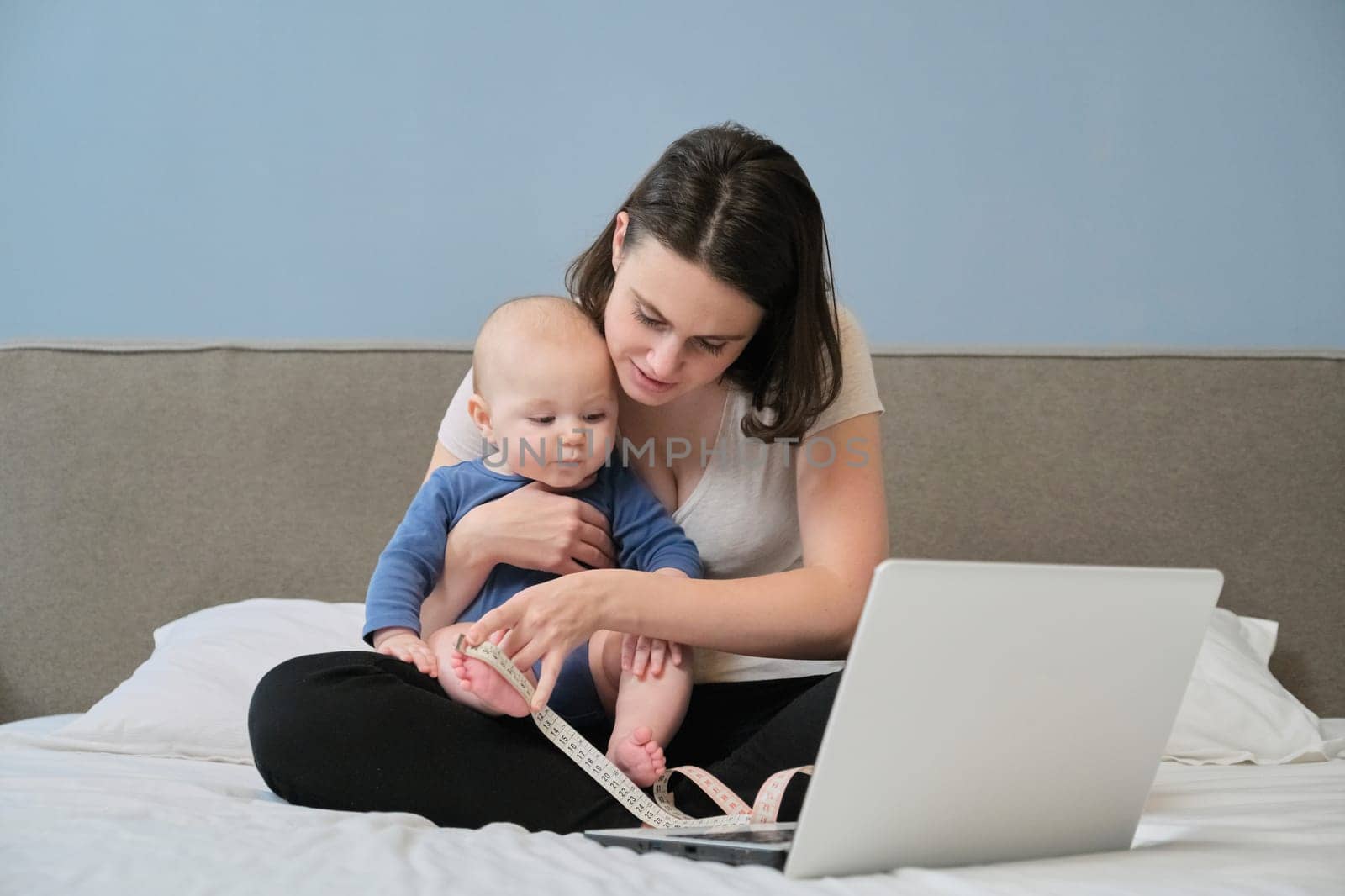 Mother with centimeter tape measuring her baby sitting at home on bed with laptop computer