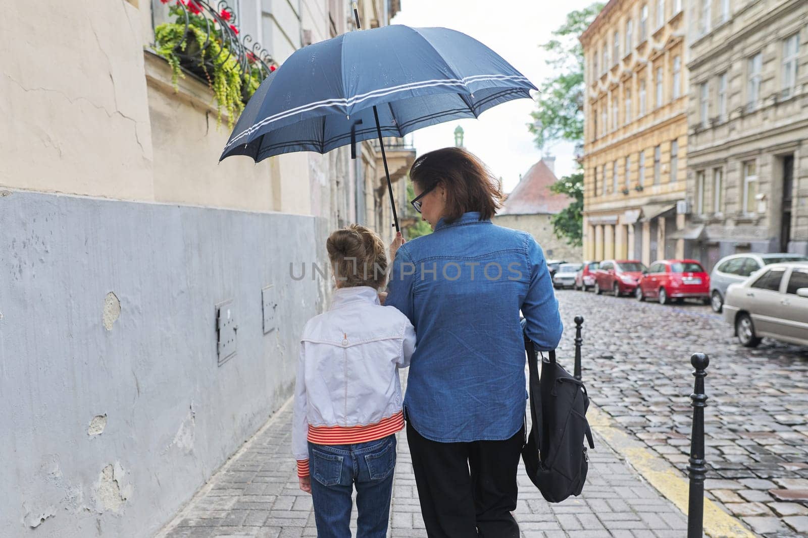 Rainy autumn weather in city, woman with child girl walking under an umbrella in street, rear view