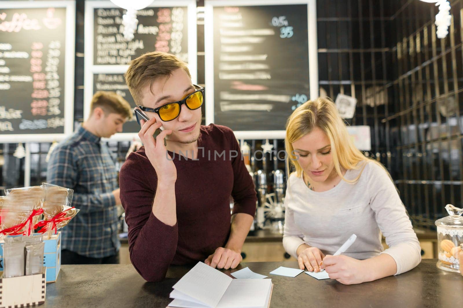 Young people working in coffee shop, man and woman barista near bar counter, receiving order by phone, background coffee machine. Teamwork, staff, small business, people concept.