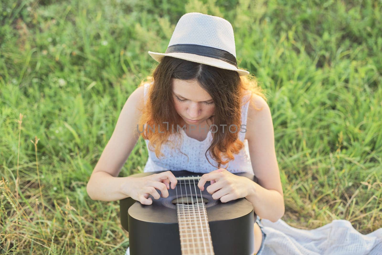 Girl teenager in hat playing guitar sitting on the grass.