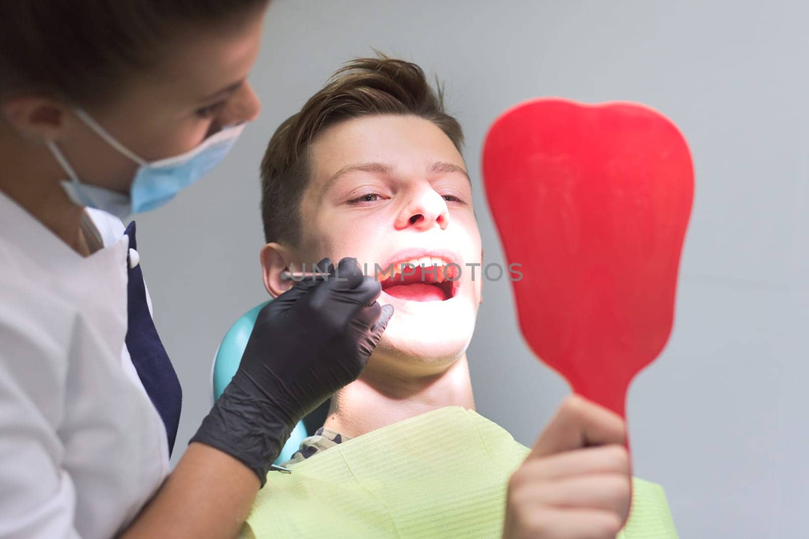 Female dentist doctor treats teeth to teenage boy sitting in dental chair looking at teeth in mirror