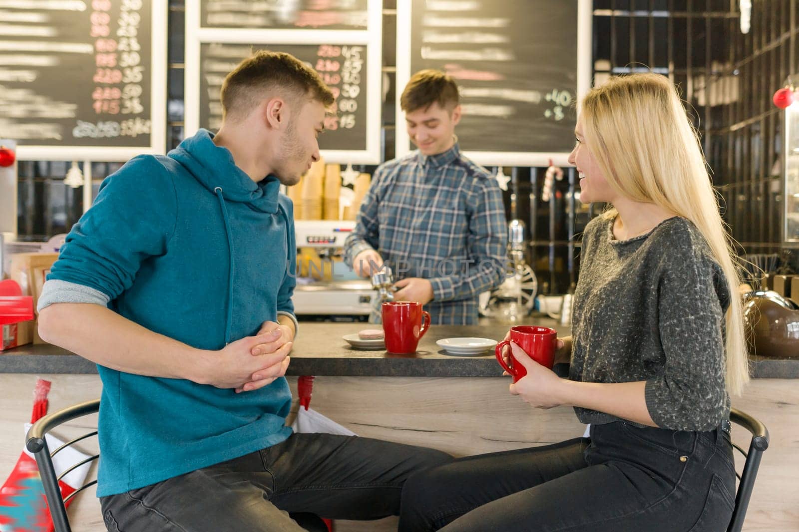 Young smiling man and woman together talking in coffee shop sitting near bar counter, couple of friends drinking tea, coffee
