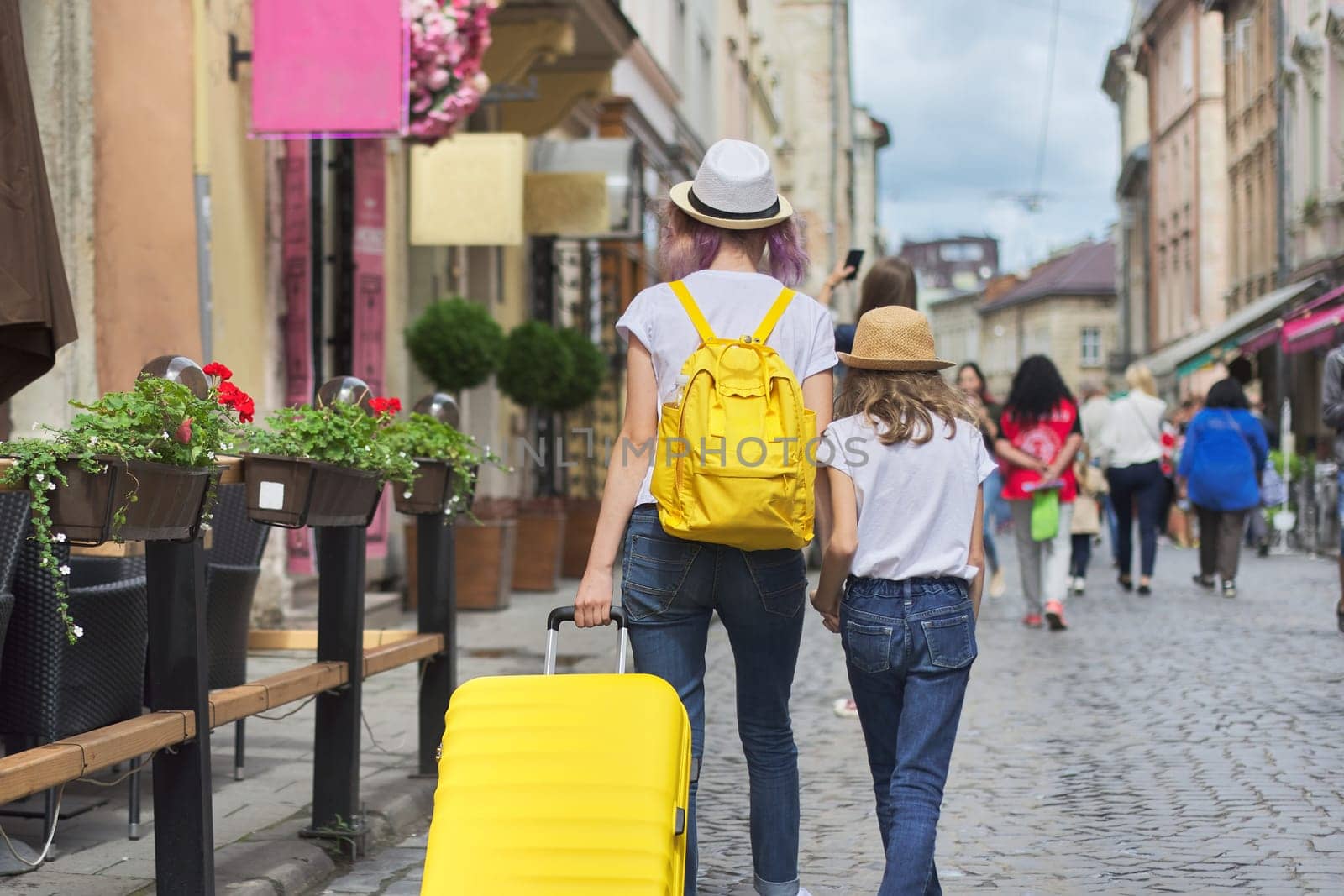 Two girls walking in tourist city holding hands with yellow suitcase, back view