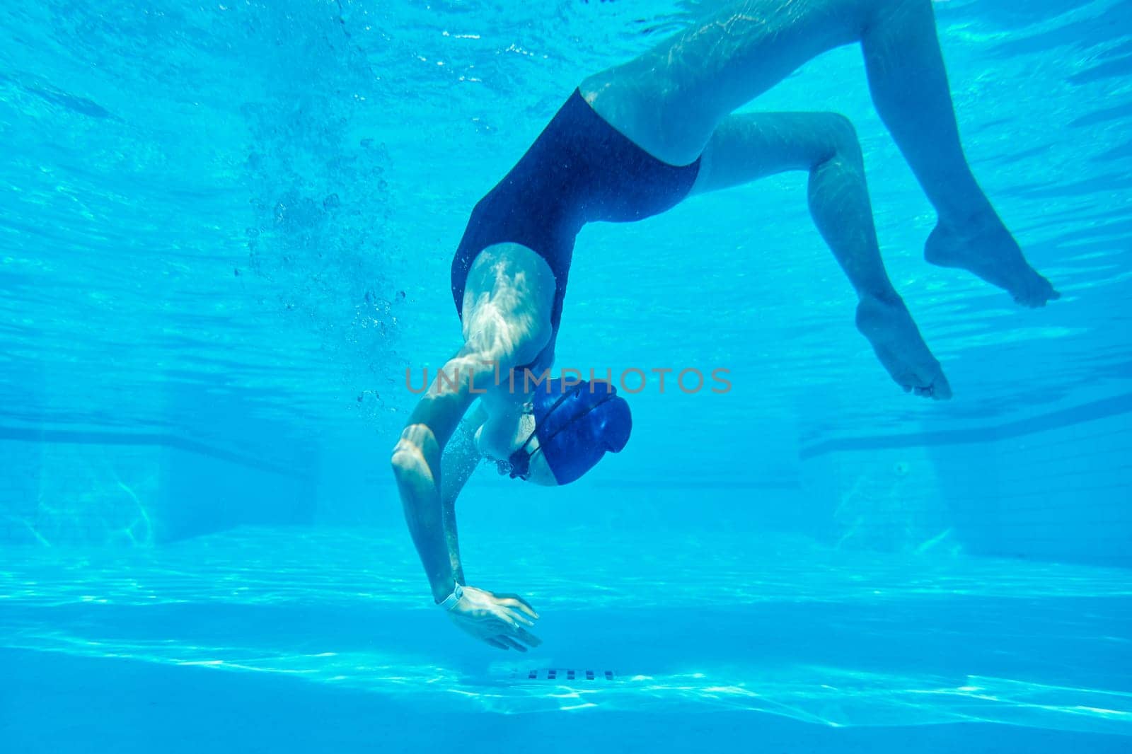 Young girl swimmer in swimsuit with goggles and swimming cap underwater in pool, female swimming under water