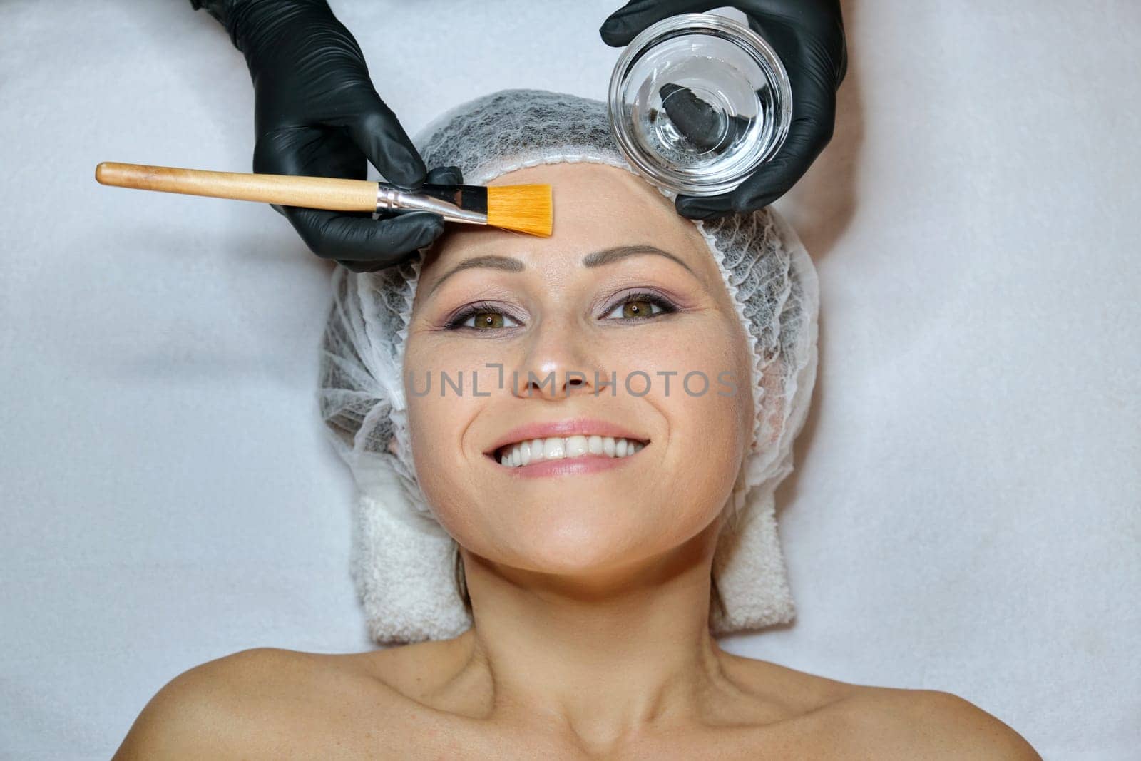 Close up of beautiful smiling happy mature woman lying on procedure in spa salon, cosmetologist applying anti-aging cosmetic by brush. People, beauty, cosmetology and skincare concept