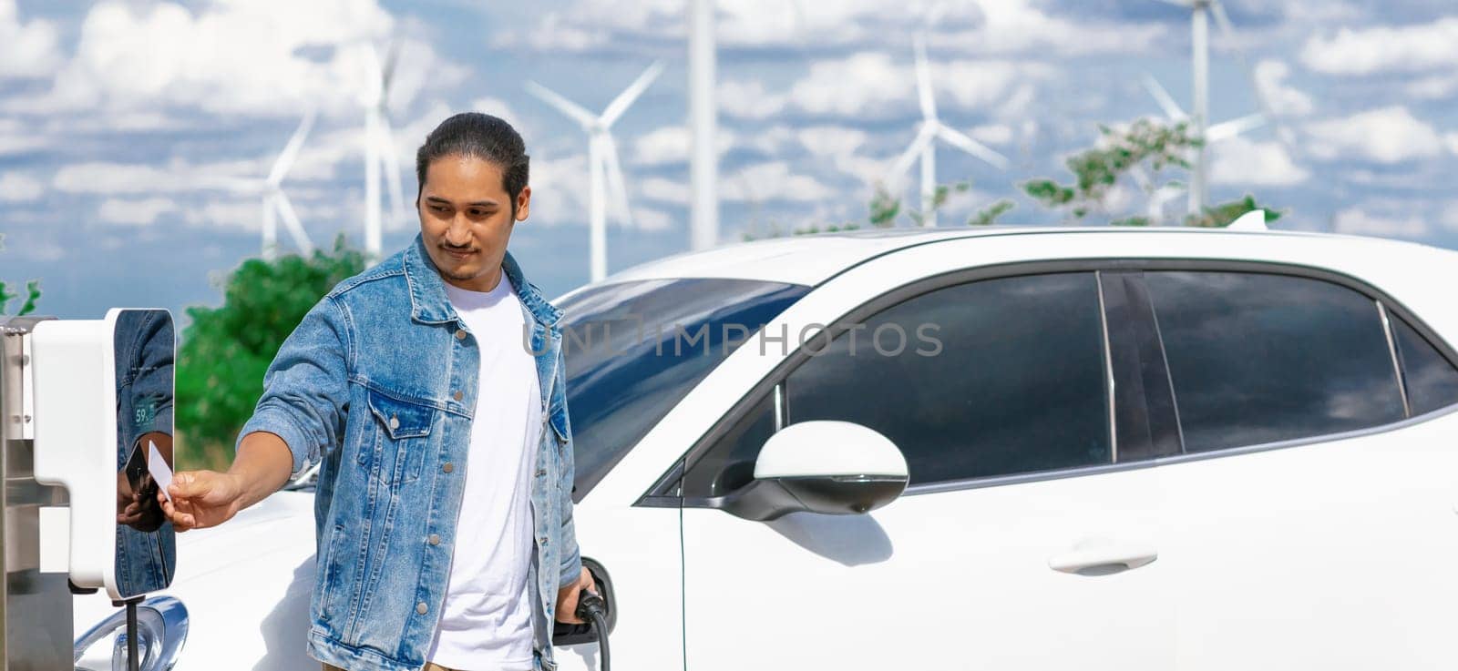 Progressive man with his electric car, EV car recharging energy from charging station on green field with wind turbine as concept of future sustainable energy. Electric vehicle with energy generator.