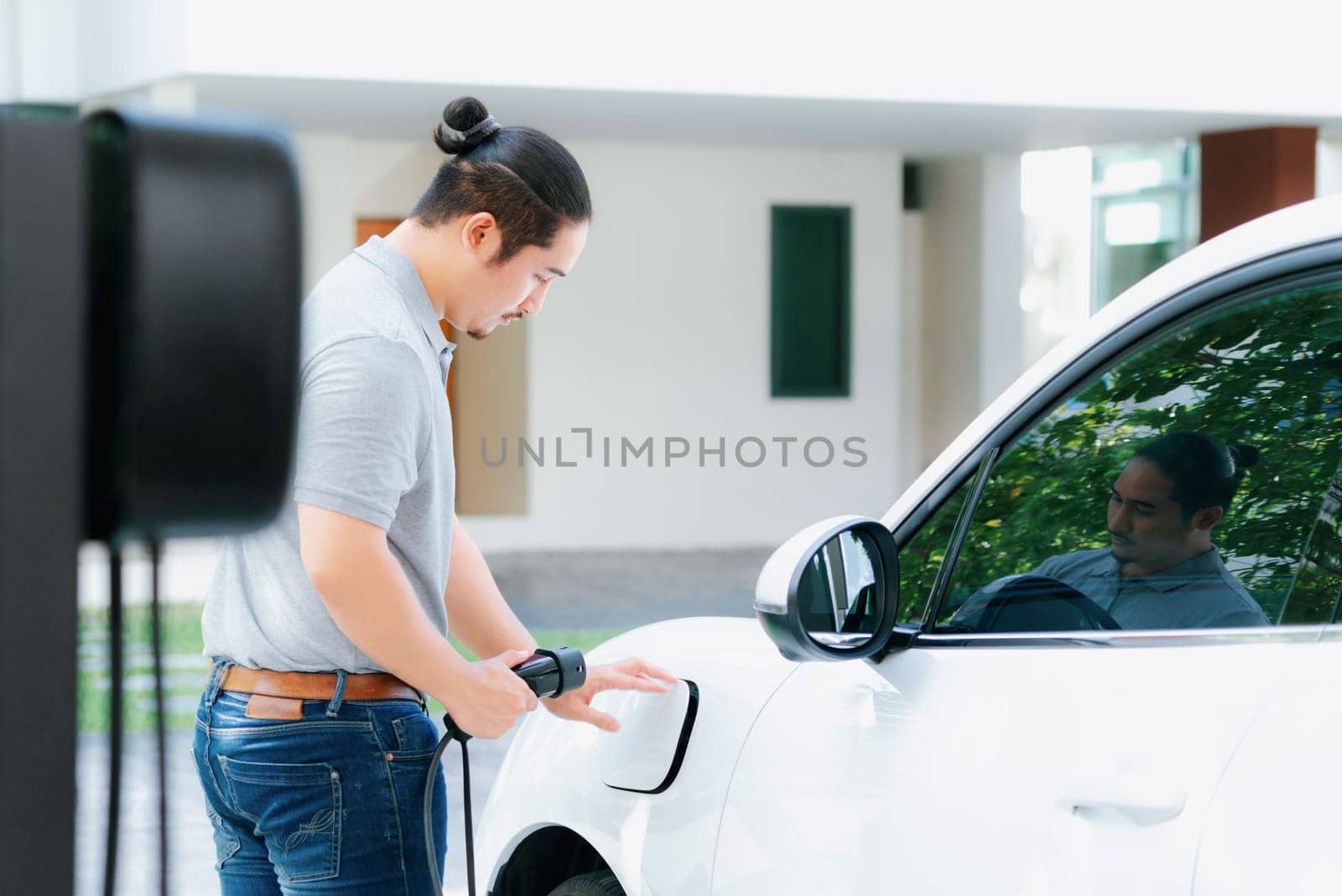 Progressive asian man install cable plug to his electric car with home charging station in the backyard. Concept use of electric vehicles in a progressive lifestyle contributes to clean environment.