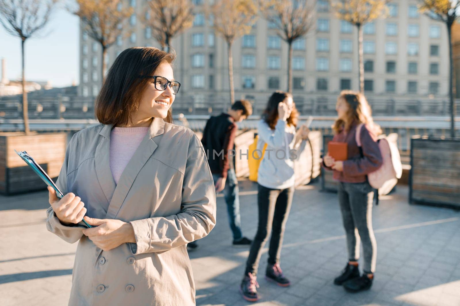 Portrait of mature smiling female teacher in glasses with clipboard, outdoor with a group of teenagers students, golden hour.