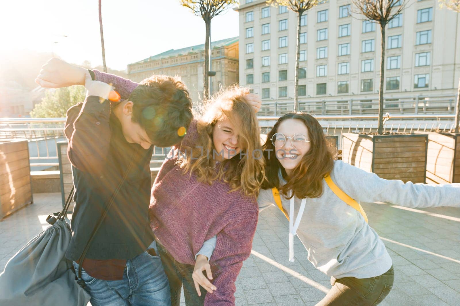 Friends teenagers students with school bags, having fun on the way from school. City background, golden hour.