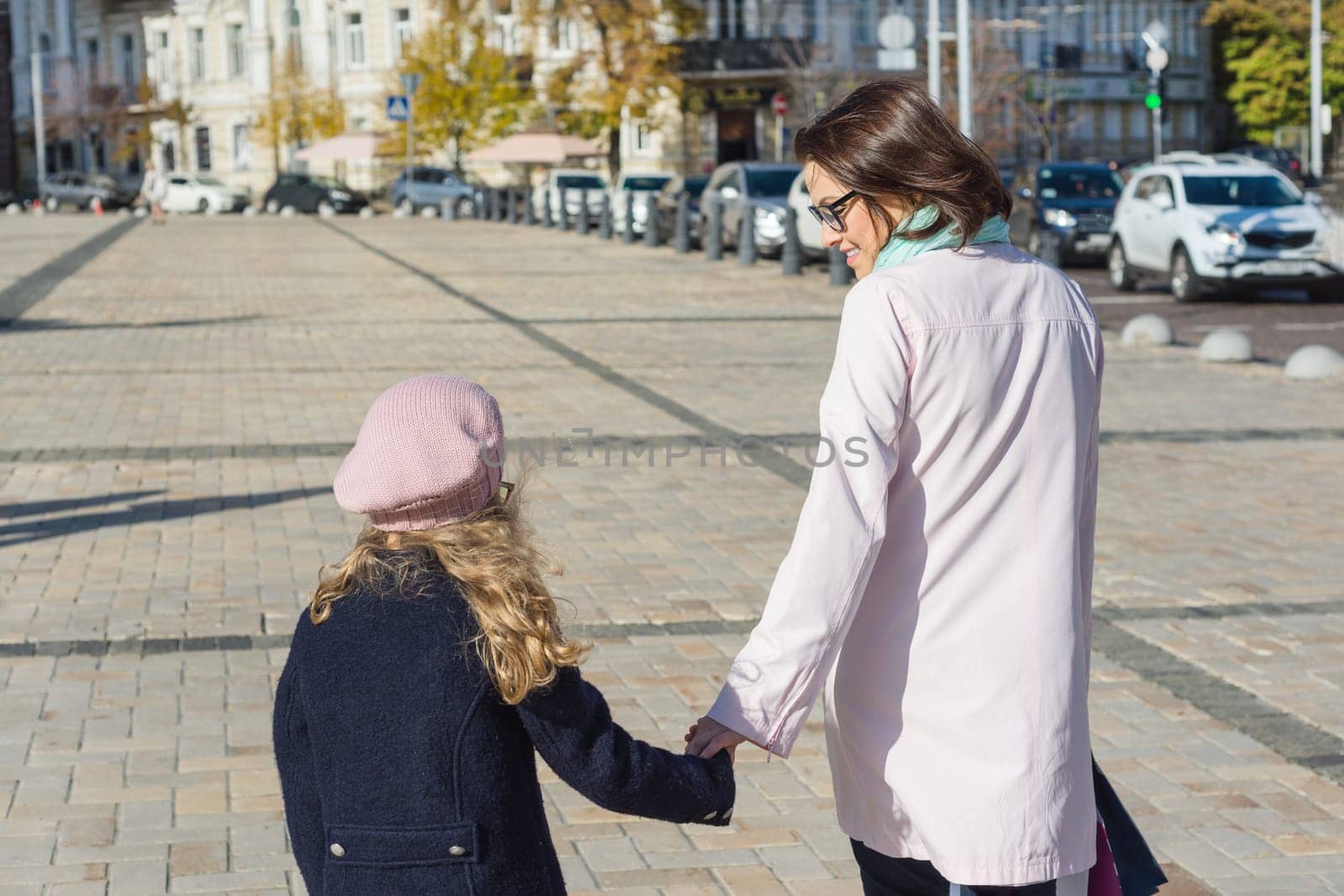Mother and daughter child hold hands, walk and talk on the city street, view from the back.