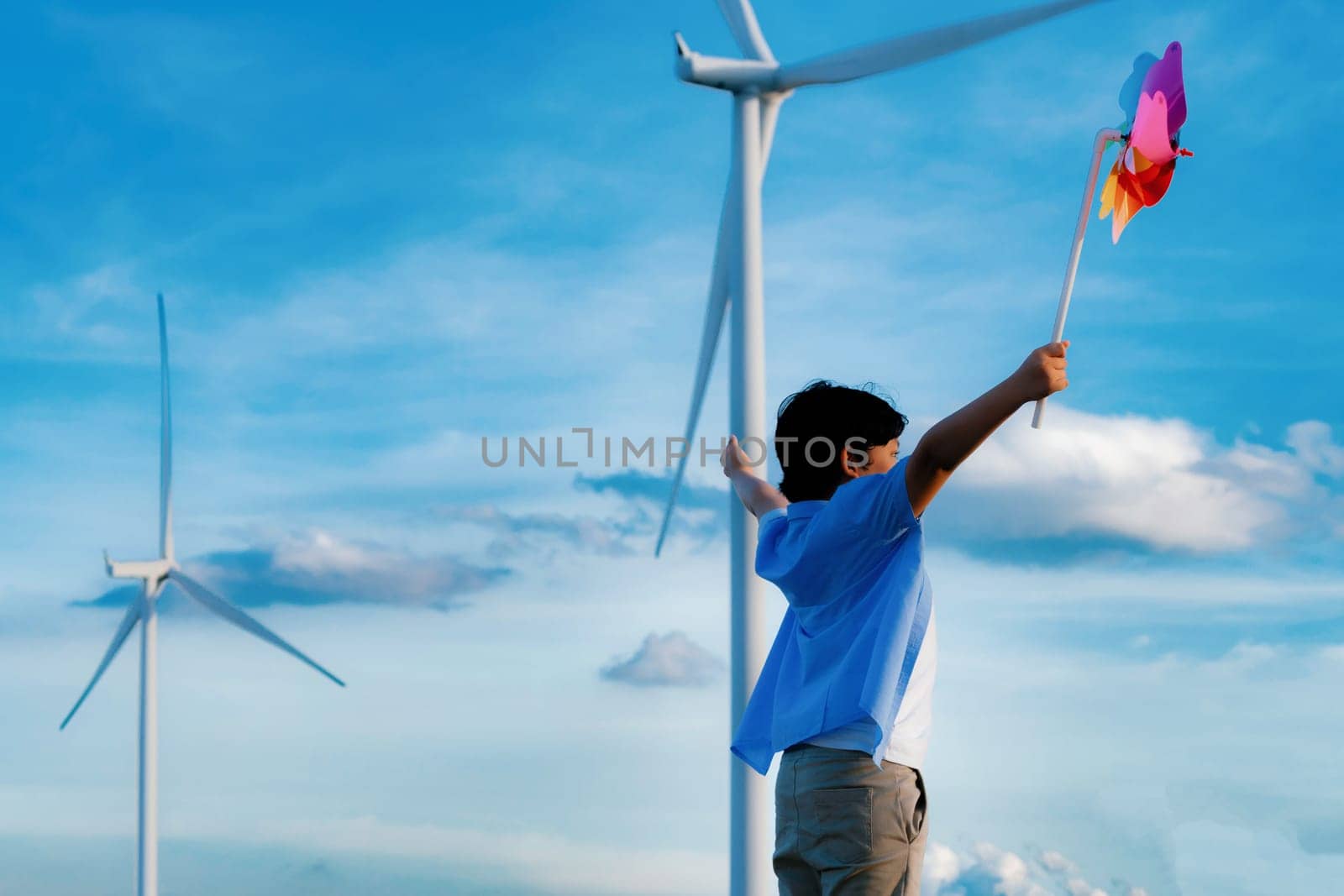 Progressive young asian boy playing with wind pinwheel toy in the wind turbine farm, green field over the hill. Green energy from renewable electric wind generator. Windmill in the countryside concept