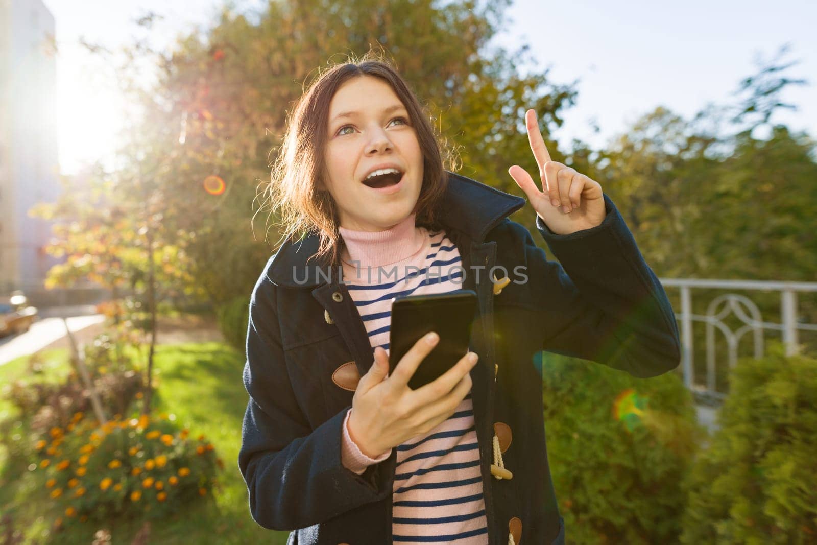 Young smiling teen girl shows index finger up, attention idea eureka, outdoor background, golden hour by VH-studio