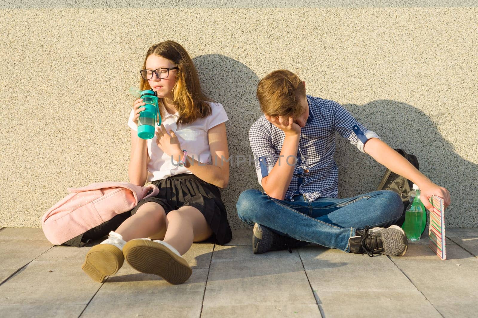 Tired teenagers schoolchildren sit near the gray wall of the school.