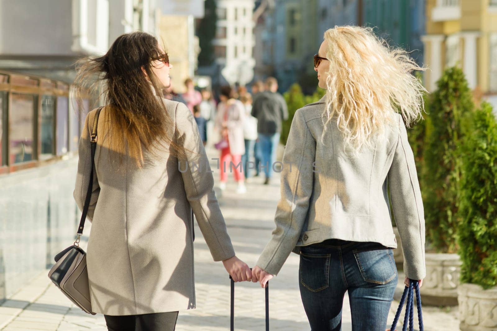 Two young women are walking along a city street, sunny autumn day, golden hour, view from the back by VH-studio